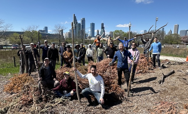A group of people stand outside smiling and holding branches, with a sunny city skyline behind them