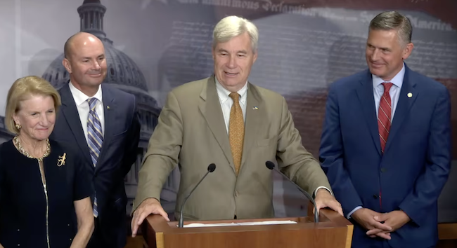 An older white man in an olive suit stands at a podium. Two men and a woman in businesswear stand around him. Everyone is smiling.
